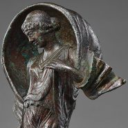Ancient Roman bronze statuette of Luna, poised on pointed toes with a windblown mantle arched over her head, holding a small object in her right hand.